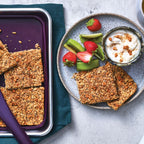 Granola Baked Apple Sweet Dip Mix Epicure bars with a dark purple spatula on a baking pan, served on a plate with yogurt, kiwi, and strawberries, with a jar of baked apples granola visible in the corner.