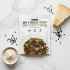 A package of Epicure Beef & Broccoli Stir-Fry is displayed on a white marble flat-lay surface, surrounded by its ingredients: a wedge of Parmesan cheese, scattered black peppercorns, and a small bowl of rice.