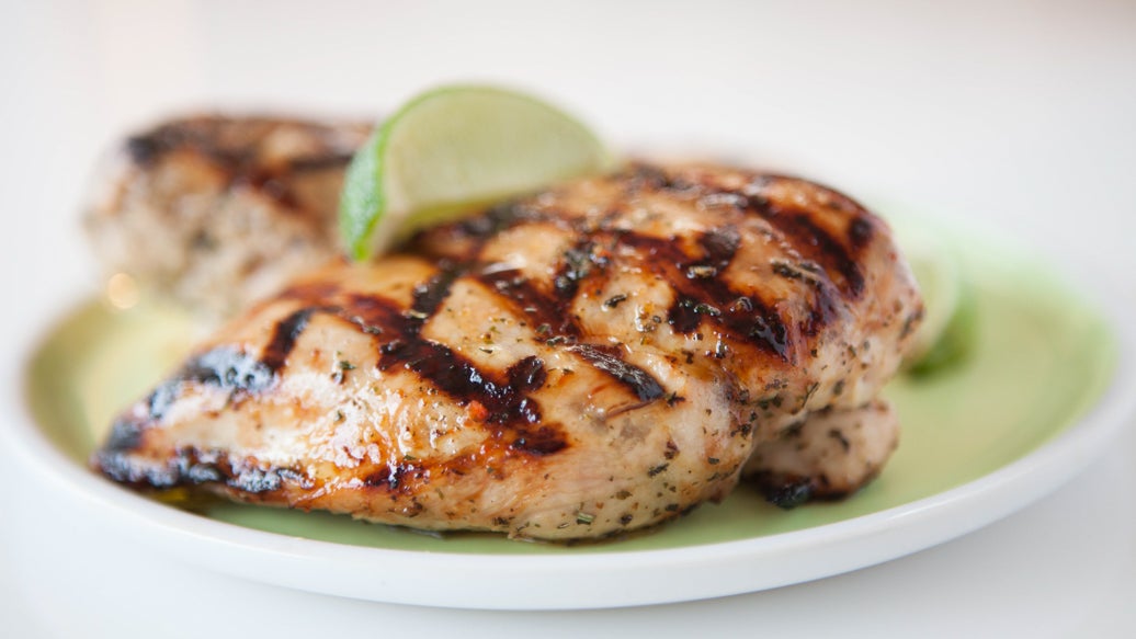 Grilled chicken breast with dark char marks on a light green plate, garnished with lime wedges, on a white plate against a bright white background.