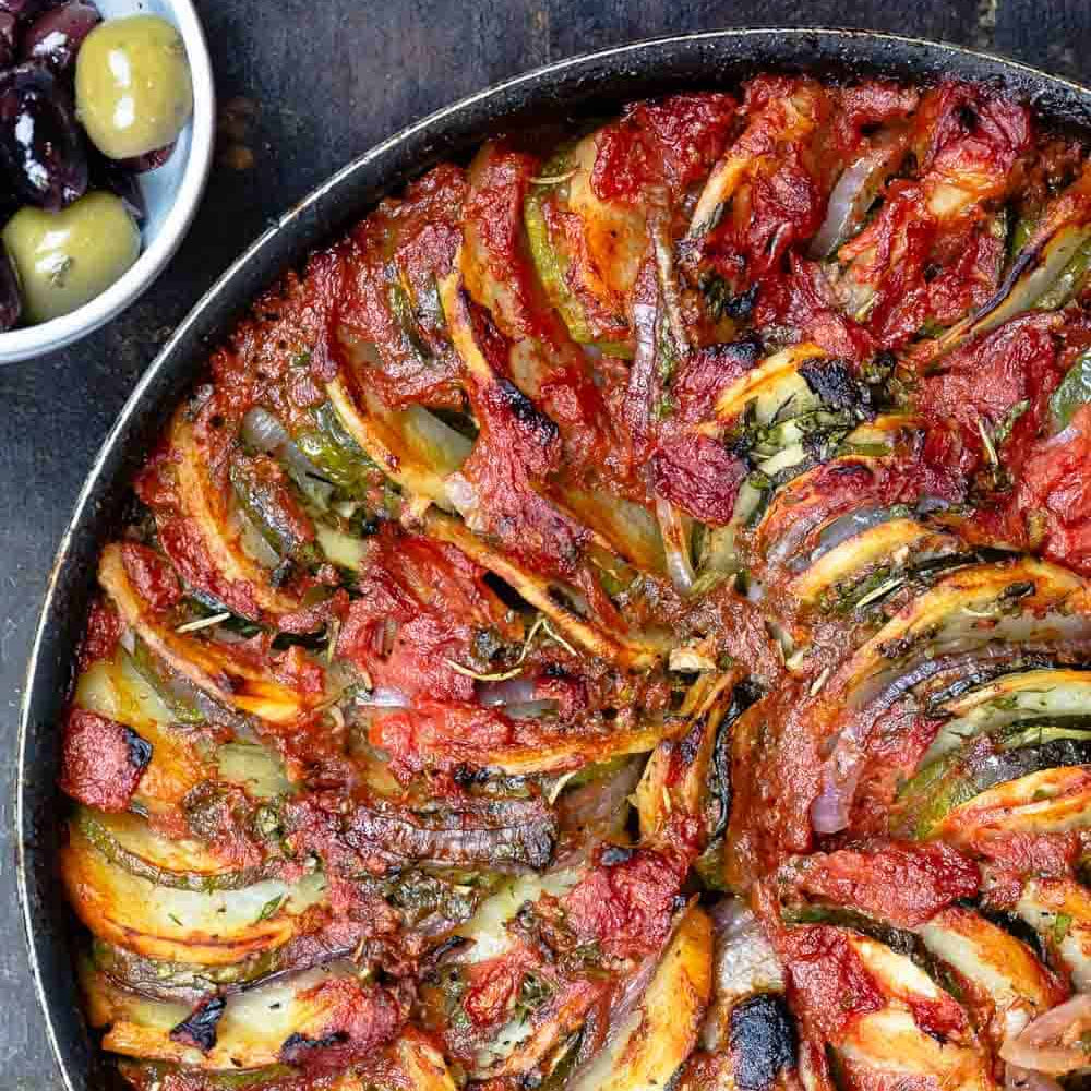 A top-down view of a round black skillet filled with baked stuffed grape leaves layered with sliced zucchini, onions, and a rich red tomato sauce, garnished with herbs.