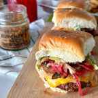A close-up of a gourmet burger on a wooden board, featuring a toasted bun, melted cheese, sautéed mushrooms, pickled red onions, and a creamy sauce. A jar of 'Butcher's Secret' is visible in the background.