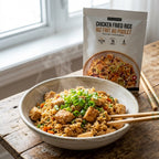 A bowl of prepared chicken fried rice with green onions sits on a rustic wooden table, with the Epicure pouch of Chicken Fried Rice standing behind it near a window.