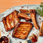 Glazed pork ribs arranged on parchment paper with a small bowl of sauce and a pastry brush on a wooden surface.