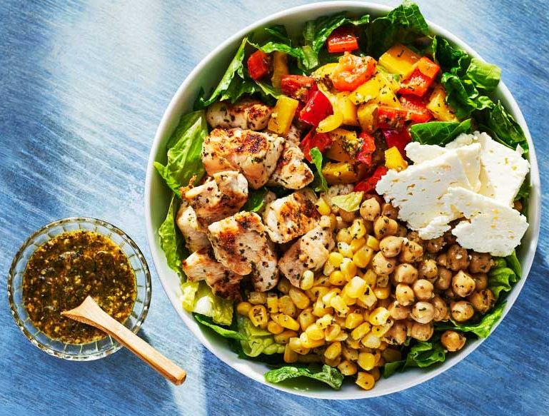 A top-down view of a large white bowl filled with a colorful salad, including greens, grilled chicken, red and yellow peppers, corn, chickpeas, and white cheese, served on a textured blue surface with a small side of Chop Chop Salad dressing.