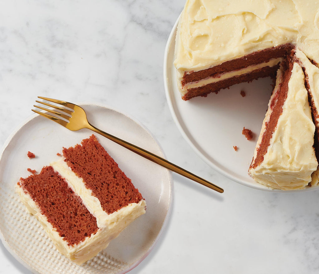 An Epicure Ruby Red Velvet cake with creamy white frosting, shown with a slice on a small plate and a gold fork, alongside the whole cake on a white plate against a light marble background.