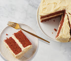 An Epicure Ruby Red Velvet cake with creamy white frosting, shown with a slice on a small plate and a gold fork, alongside the whole cake on a white plate against a light marble background.