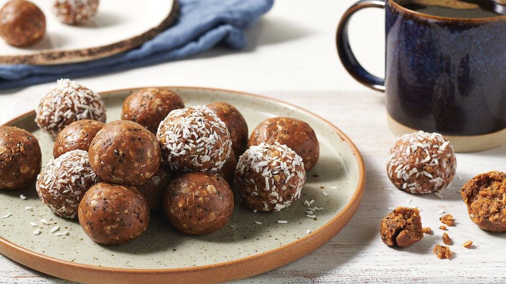 A plate of brown energy balls, some rolled in coconut flakes, with a dark ceramic mug and a few loose balls on a light wooden table.