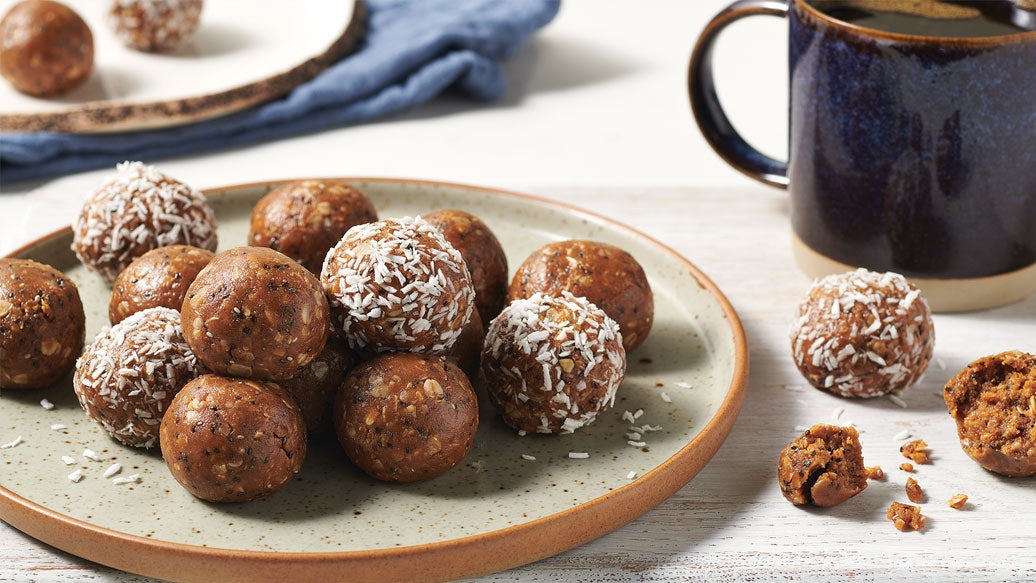 A plate of brown energy balls, some rolled in coconut flakes, with a dark ceramic mug and a few loose balls on a light wooden table.