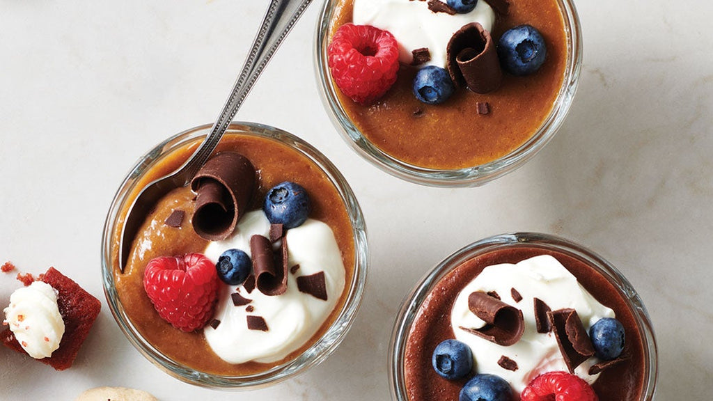 Three small glass jars of Epicure Dulce de Leche Pudding topped with whipped cream, blueberries, raspberries, and chocolate curls, arranged on a light marble surface with a spoon in one jar.