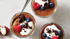 Three small glass jars of Epicure Dulce de Leche Pudding topped with whipped cream, blueberries, raspberries, and chocolate curls, arranged on a light marble surface with a spoon in one jar.