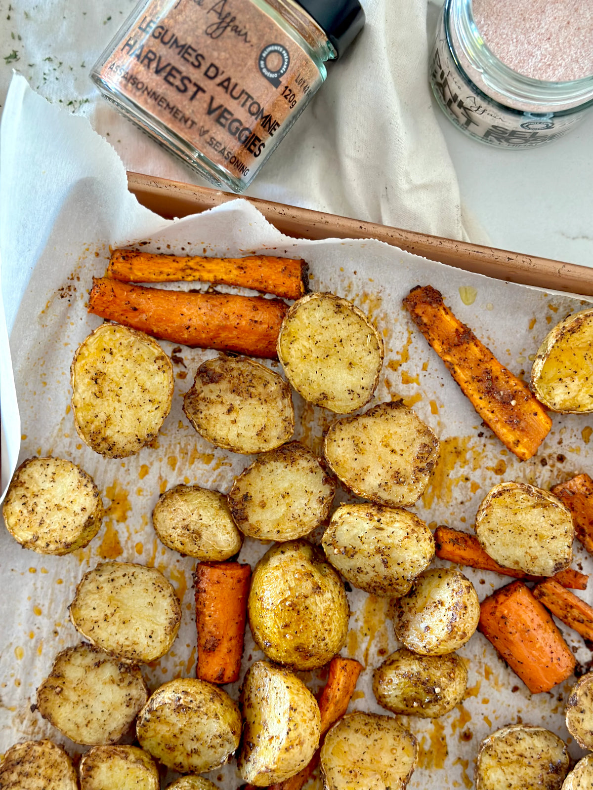 Overhead view of roasted baby potatoes and sweet potato sticks on a parchment-lined baking sheet, with seasoning jars partially visible at the top.