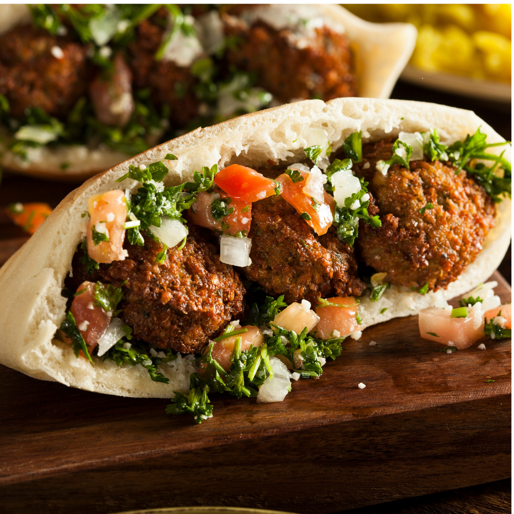 A close-up of a stuffed pita bread sandwich filled with browned falafels, chopped parsley, diced tomatoes, and onions, served on a dark wooden board.