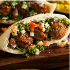 A close-up of a stuffed pita bread sandwich filled with browned falafels, chopped parsley, diced tomatoes, and onions, served on a dark wooden board.