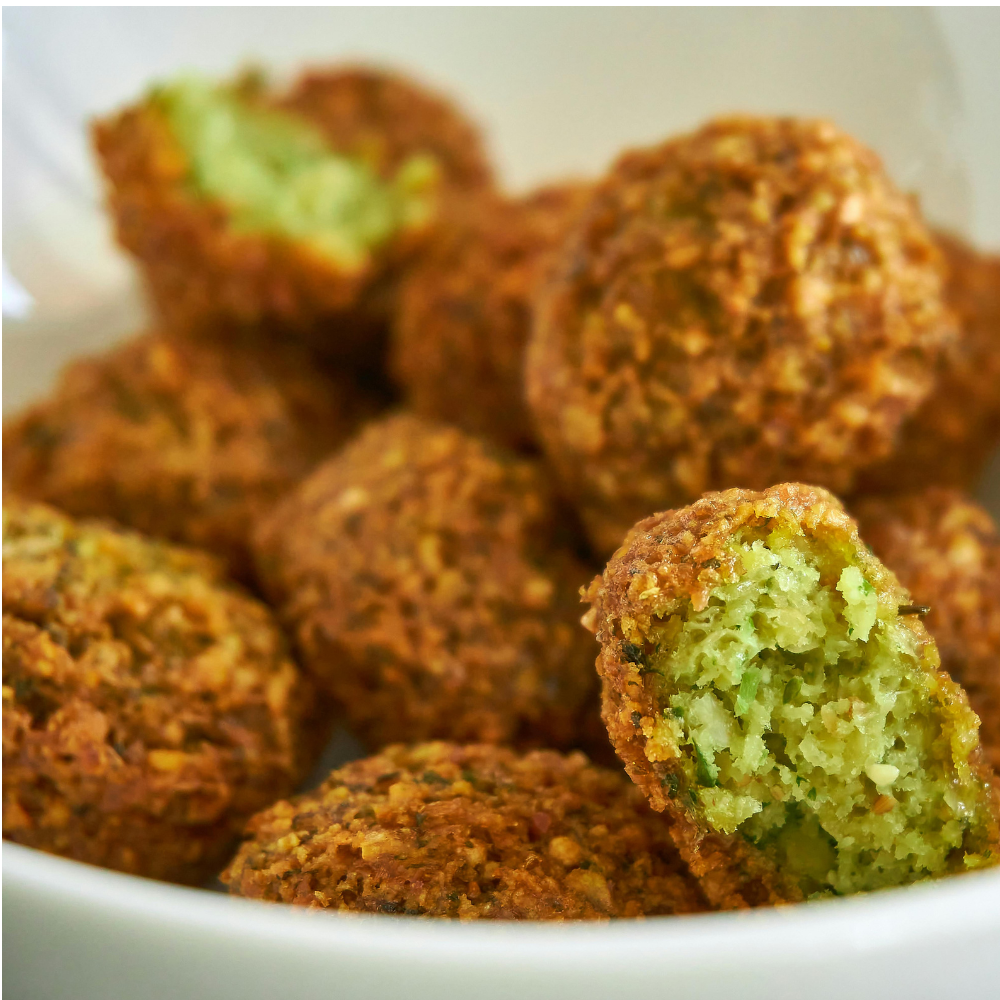 Close-up of golden-brown fried falafel balls in a white bowl, with one ball broken open to show a green herb filling.