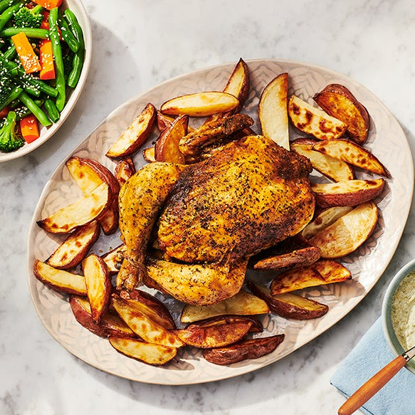 A roasted whole chicken with golden-brown skin seasoned with Feisty Green Sauce Mix by Epicure sits on a large oval platter surrounded by roasted potato wedges, styled on a light marble surface with a side salad, dip, plates, and a glass of water.