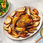 A roasted whole chicken with golden-brown skin seasoned with Feisty Green Sauce Mix by Epicure sits on a large oval platter surrounded by roasted potato wedges, styled on a light marble surface with a side salad, dip, plates, and a glass of water.
