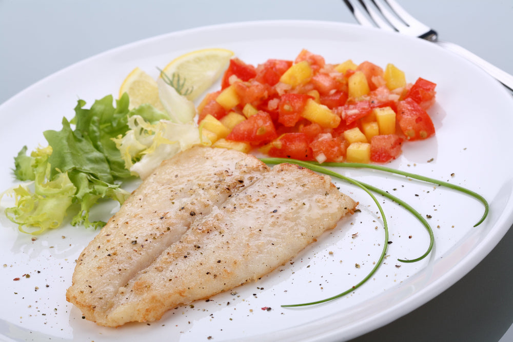 Pan fried Whitefish fillet seasoned with Fish seasoning on a white plate accompanied by salsa, lemon slices and green leaves 