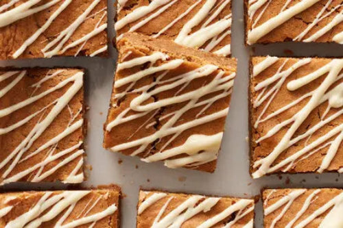 Close-up of several Gingerbread Blondies arranged on a baking sheet, with one bar lifted slightly to show the texture. The bars are topped with white chocolate drizzle.