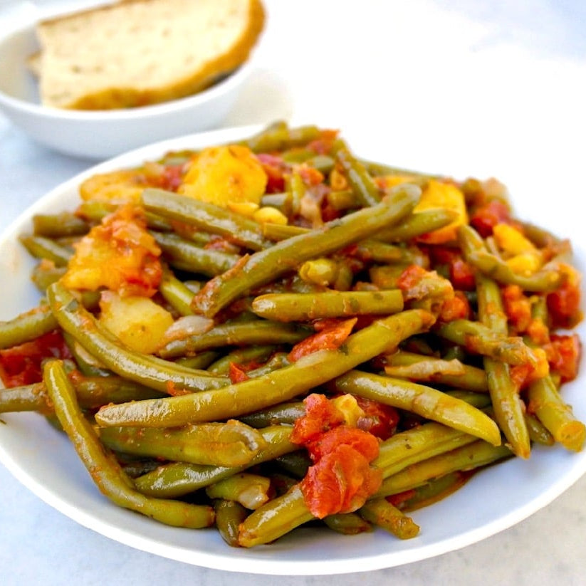 A white plate of cooked green beans with diced tomatoes and yellow potato pieces, served on a light surface with a small bowl of bread in the background.