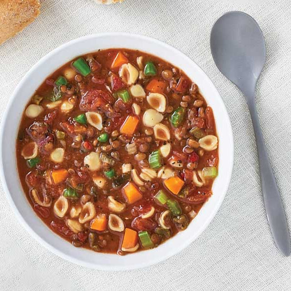 A top-down view of a white bowl filled with minestrone soup, featuring small pasta shells, carrots, green beans, and lentils, set on a textured white tablecloth next to a gray spoon.
