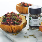 A jar of Epicure Holiday Fétes seasoning displayed on a white marble board with halved roasted squash topped with a colorful herb and seed mixture, on a clean white background.