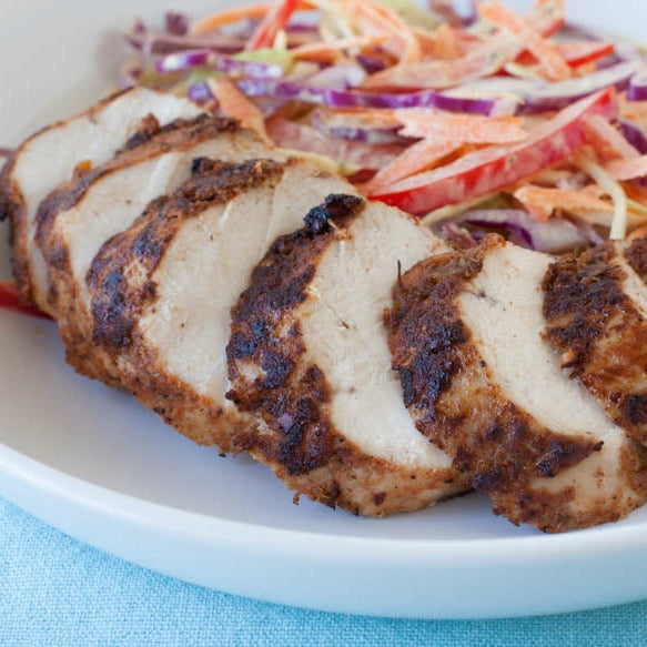 Grilled Jerk chicken breast slices served on a white plate with a colorful slaw of shredded carrots, red cabbage, and white cabbage on a light blue textured tablecloth.