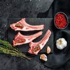 Three raw bone-in lamb chops with visible fat marbling, arranged on a dark stone serving board with herbs, garlic cloves, and a small bowl of sumac berries on a dark textured surface.