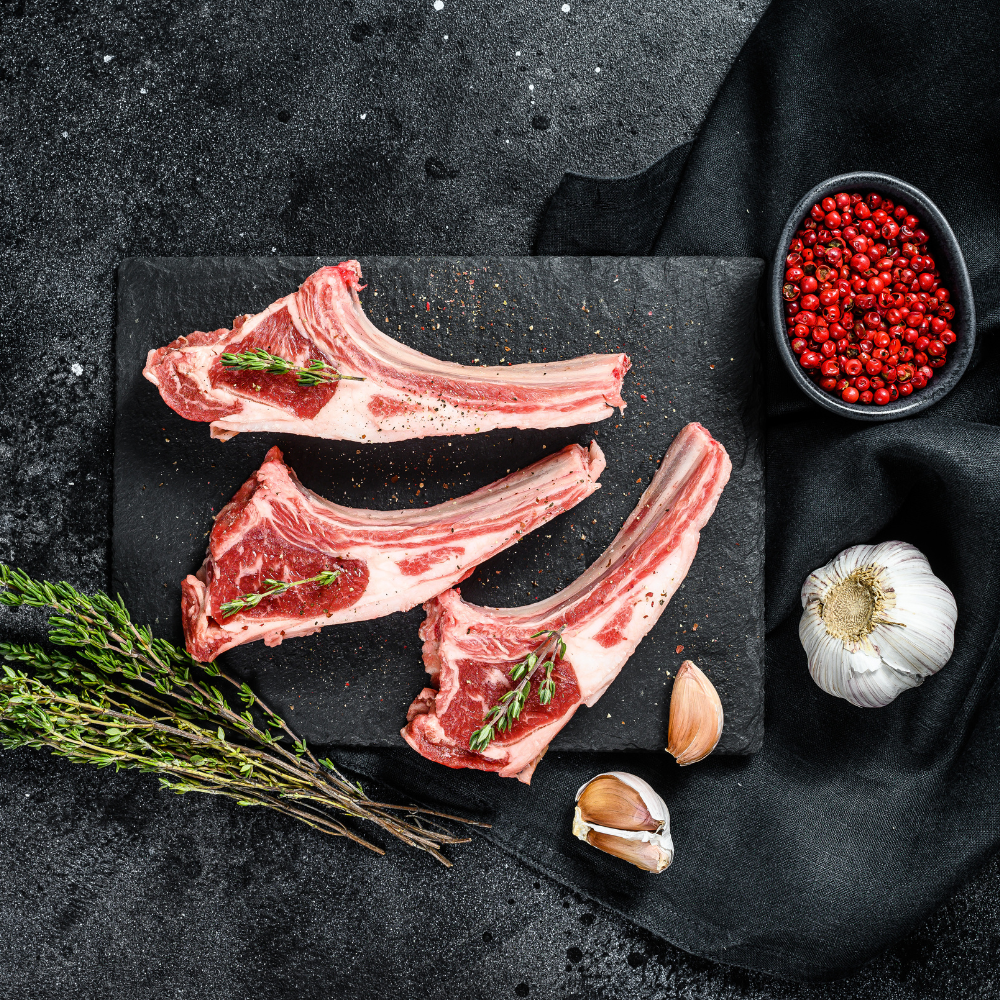 Three raw bone-in lamb chops with visible fat marbling, arranged on a dark stone serving board with herbs, garlic cloves, and a small bowl of sumac berries on a dark textured surface.