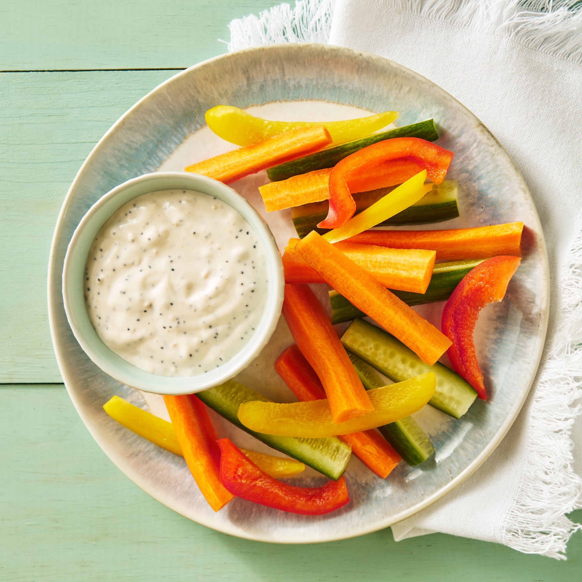 A top-down view of a round ceramic plate filled with colorful carrot sticks, cucumber sticks, red bell pepper strips, and yellow bell pepper strips, served with a small bowl of Lemon Poppy Dip on a light green wooden table with a white fringed napkin.