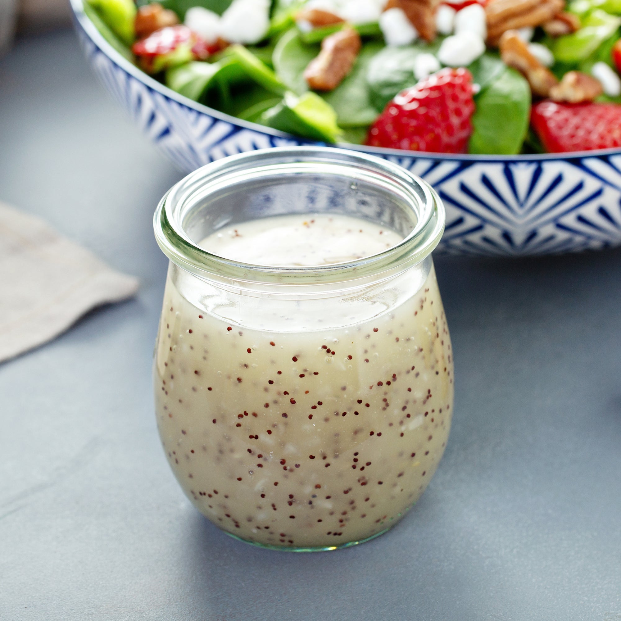 A small clear glass jar filled with a Lemon Poppy Seed dressing, set on a gray surface with a blurred salad bowl in the background.