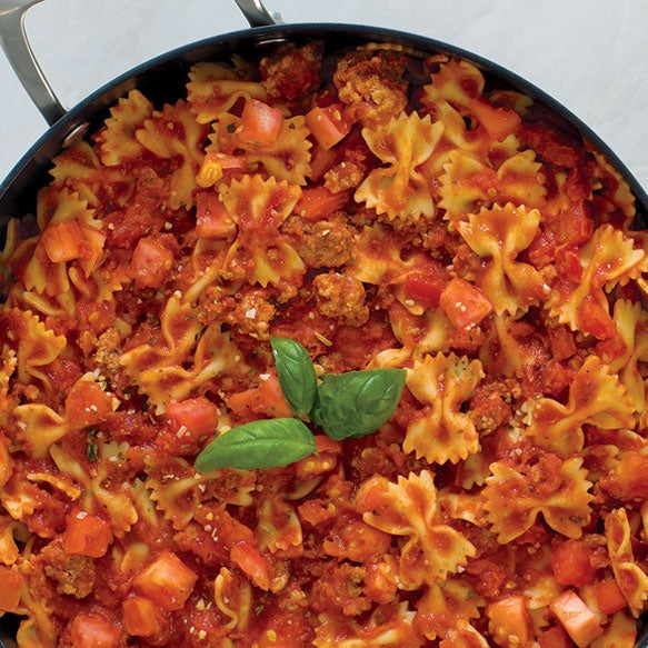 A top-down view of a black skillet filled with bowtie pasta in a tomato and meat sauce, garnished with fresh basil leaves. The pan sits on a light marble surface surrounded by culinary ingredients including garlic, tomatoes, a lemon wedge, and additional basil leaves.