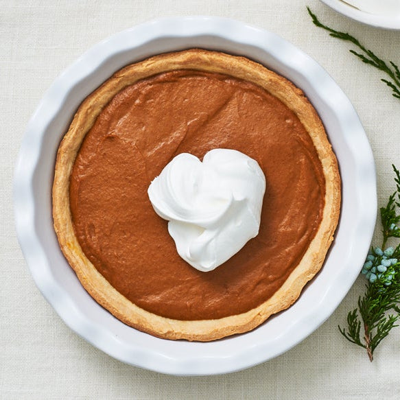 A top-down view of a pumpkin pie in a white ceramic dish, topped with a dollop of whipped cream, set on a light beige linen tablecloth with a sprig of green herbs and a gold spoon handle as props.