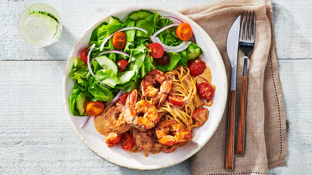 A top-down view of a white plate filled with shrimp pasta, sliced tomatoes, and a fresh green salad with cucumber and red onion, set on a light wood table with a fork and knife on a beige napkin.