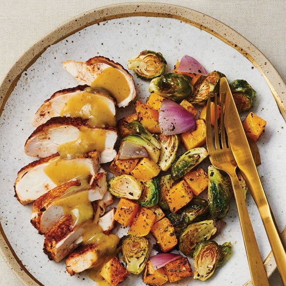 A plated prepared meal featuring sliced grilled chicken with yellow sauce, roasted Brussels sprouts, and sweet potato cubes on a speckled ceramic plate, with a side of salad and a small dish of sauce on a light beige table.