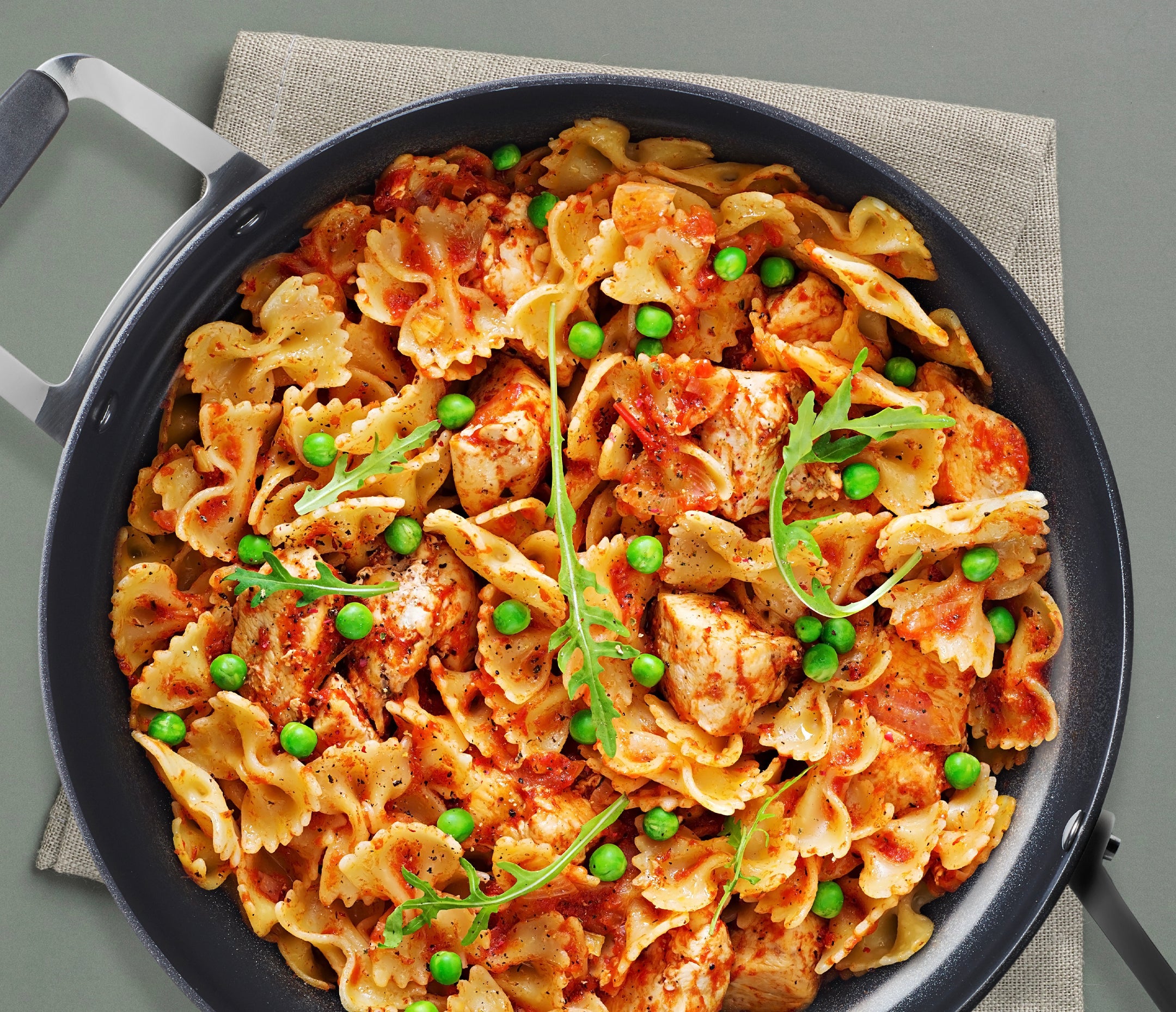 A top-down view of a black skillet filled with bowtie pasta, chicken, Epicure Rosee sauce, green peas, and arugula, set on a beige cloth over a gray surface.
