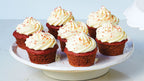 Six Epicure Ruby red velvet cupcakes with white frosting and orange sprinkles, arranged on a white ceramic cake stand against a light blue background.