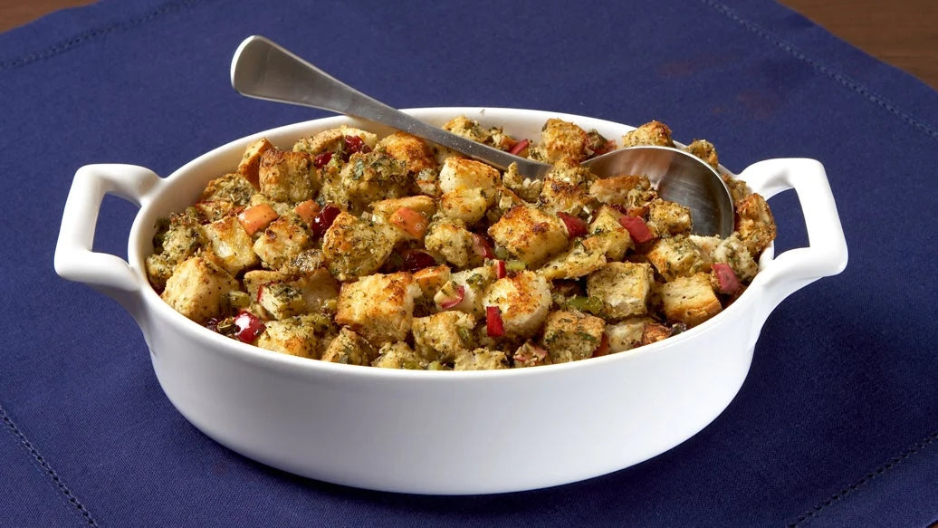 A white ceramic baking dish filled with Epicure Sage & Apple stuffing or dressing, with a metal spoon resting inside, set on a dark blue fabric surface.