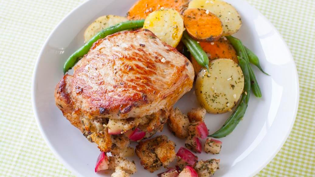 A plated prepared meal featuring a Stuffed Pork Chop with Epicure Sage & Apple Seasoning stuffing, served with roasted potatoes, green beans, and stuffing cubes on a white plate over a green-and-white checkered tablecloth.