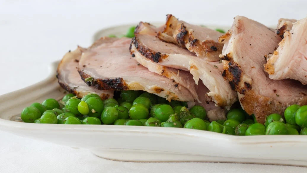 Sliced pork loin with Epicure Sage & Apple Stuffing Seasoning with charred edges served over bright green peas on a white plate against a light neutral background.