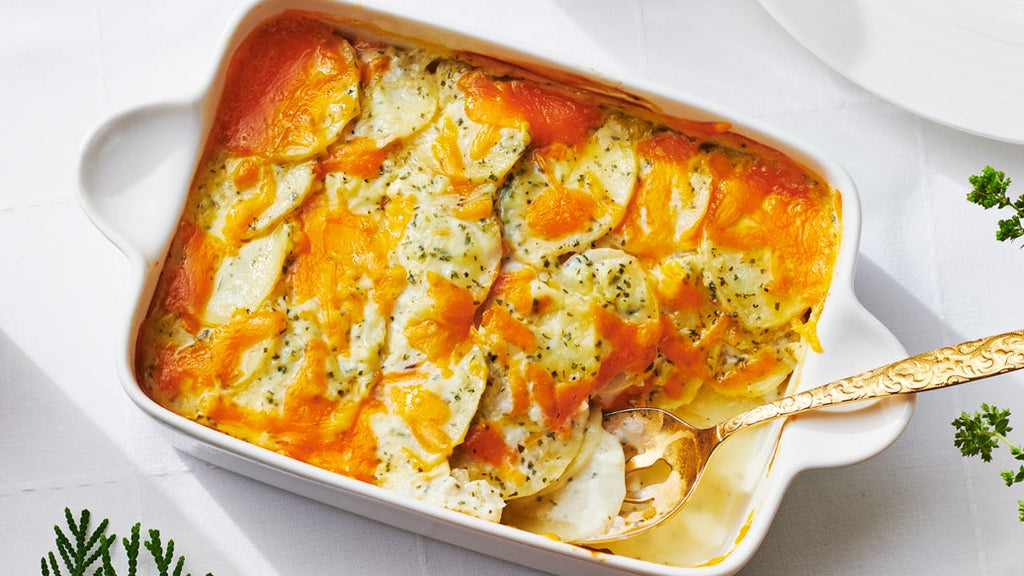 A baked Scalloped potato casserole in a white rectangular baking dish, topped with melted orange cheese, broccoli, and herbs, with a gold spoon resting in the dish on a white surface.