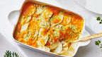 A baked Scalloped potato casserole in a white rectangular baking dish, topped with melted orange cheese, broccoli, and herbs, with a gold spoon resting in the dish on a white surface.