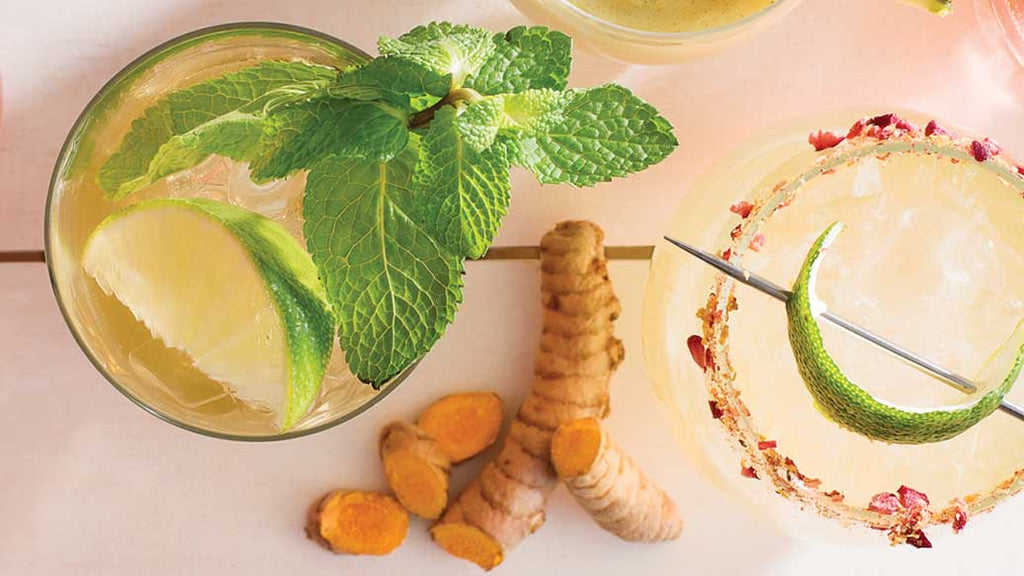 A styled flat-lay of two Sencha Jasmine Mojito cocktails on a pale pink surface. One glass contains a light yellow drink with a lime wedge and fresh mint leaves, and the other has a pale yellow drink rimmed with dried pink flowers and a lime garnish on a metal spoon.