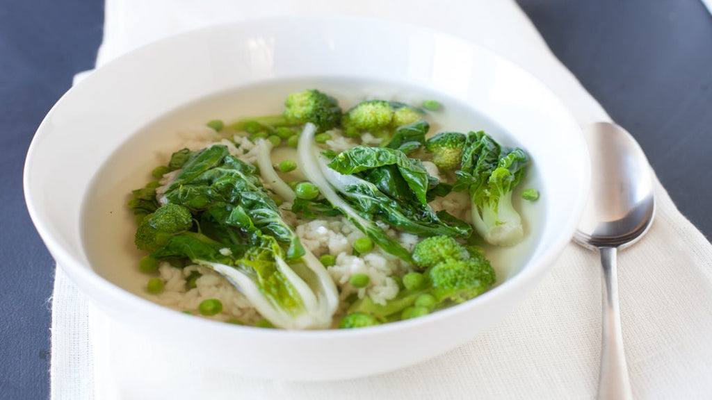 A white ceramic bowl filled with a Sencha Tea Soup containing green broccoli florets, bok choy, and green peas, served on a white napkin with a silver spoon on a dark gray surface.