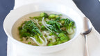 A white ceramic bowl filled with a Sencha Tea Soup containing green broccoli florets, bok choy, and green peas, served on a white napkin with a silver spoon on a dark gray surface.