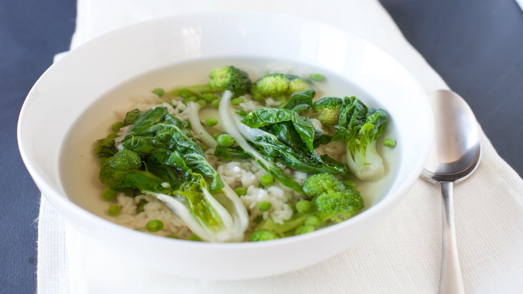 A white ceramic bowl filled with a Sencha Tea Soup containing green broccoli florets, bok choy, and green peas, served on a white napkin with a silver spoon on a dark gray surface.