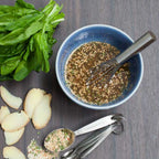 Sliced ginger, measuring spoons with mixed herbs, and a small piece of ginger root. arranged on a dark wood surface: a bunch of fresh green spinach leaves, a blue bowl with a whisk containing a Prepared Epicure Sesame Ginger Dressing.