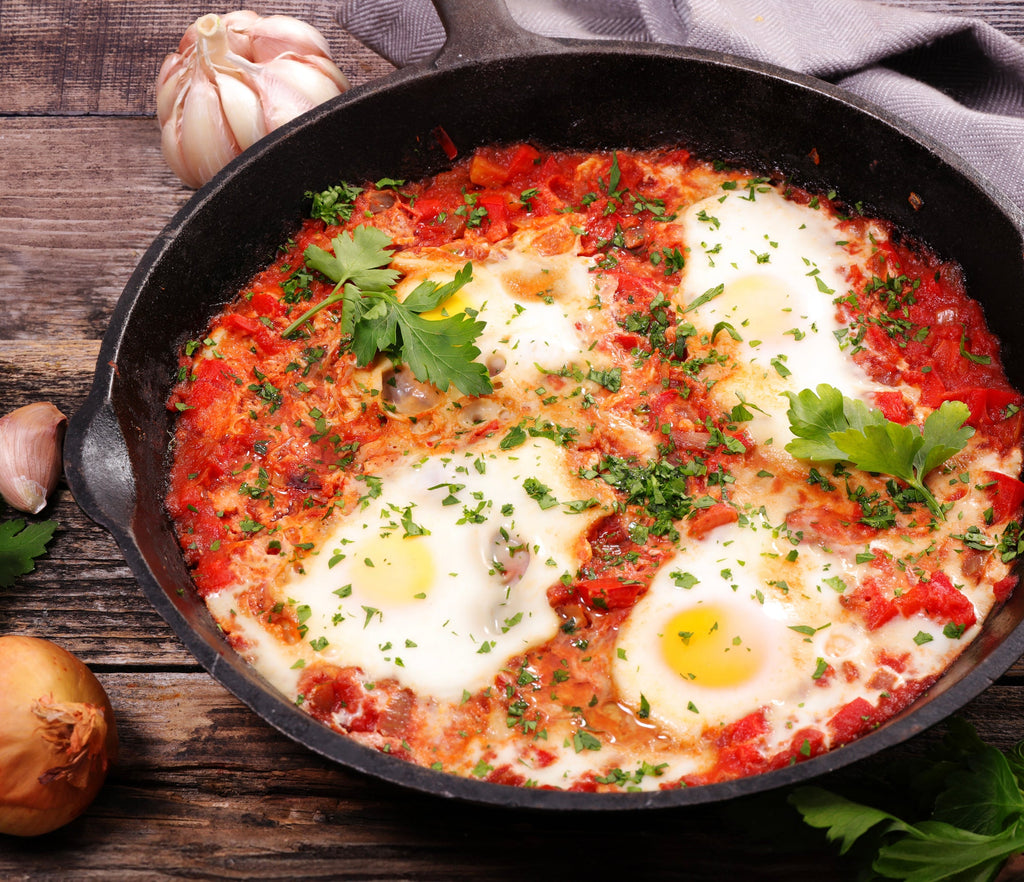 A cast iron skillet filled with shakshuka, featuring sunny-side-up eggs in Epicure Rosee sauce, garnished with chopped parsley, set on a rustic wooden table with garlic and an onion nearby.