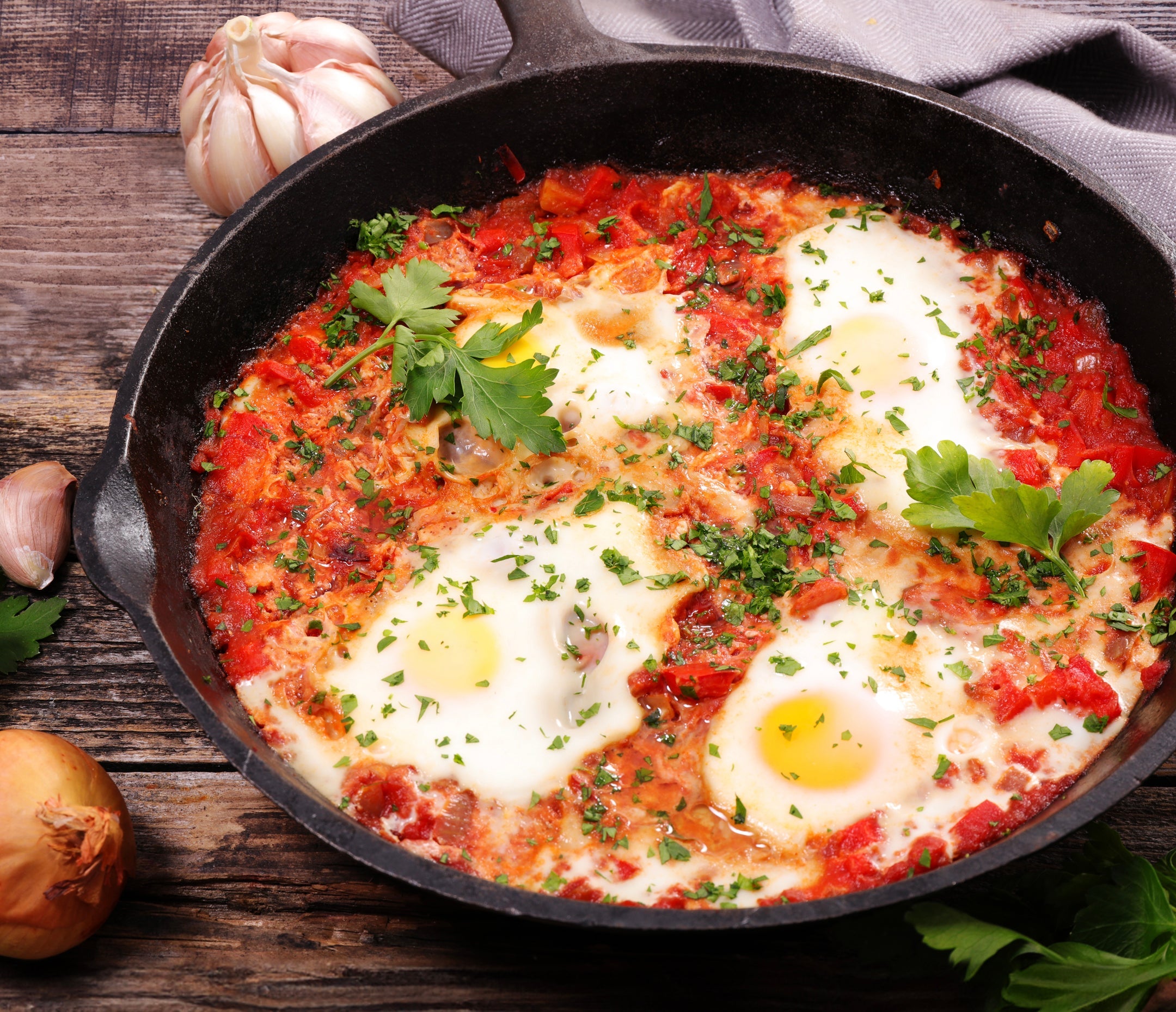 A cast iron skillet filled with shakshuka, featuring sunny-side-up eggs in Epicure Rosee sauce, garnished with chopped parsley, set on a rustic wooden table with garlic and an onion nearby.
