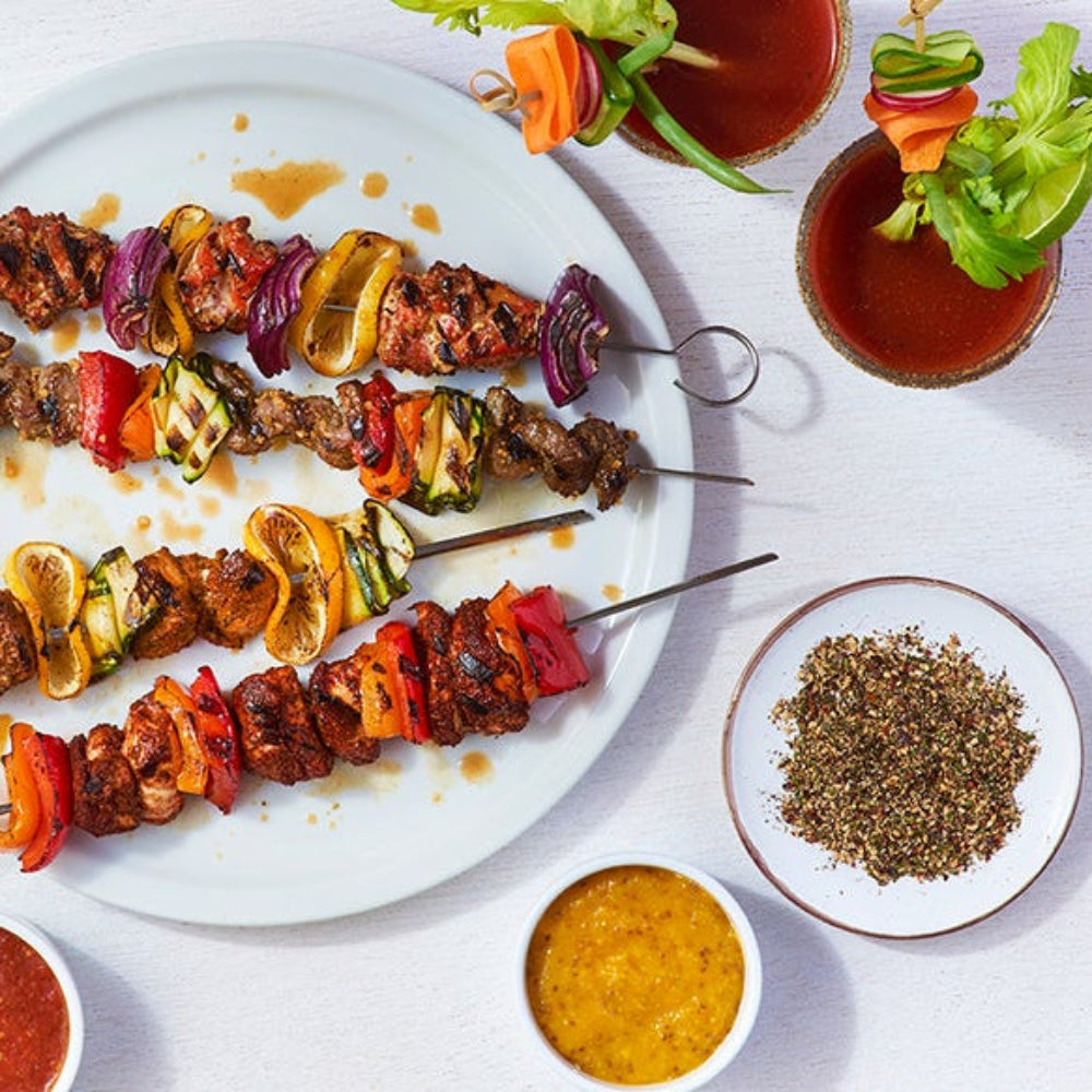 Four skewers of vegetable and meat shish kebabs on a large white plate, styled with small bowls of red and yellow dipping sauces, a bowl of Epicure Shish Kebab Seasoning, and garnished vegetables on a white background.