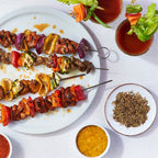 Four skewers of vegetable and meat shish kebabs on a large white plate, styled with small bowls of red and yellow dipping sauces, a bowl of Epicure Shish Kebab Seasoning, and garnished vegetables on a white background.
